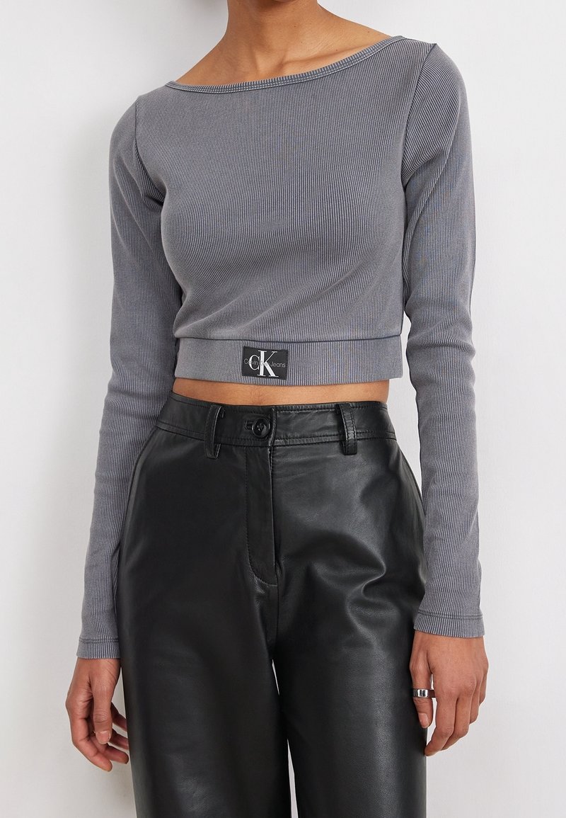 Grå ribbad långärmad croppad topp med bred halsringning, prydd med en svart CK Jeans-logotyp vid fållen, som är ihopparad med svarta high-waisted läderbyxor.