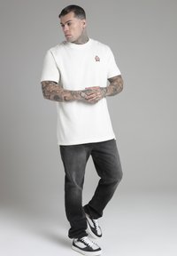 T-shirt a maniche corte color avorio chiaro con un piccolo stemma ricamato, abbinato a jeans grigi scuri e sneaker bianco e nere. Tessuto di cotone liscio.