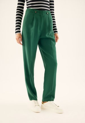 Person trägt grüne, hoch taillierte Hose mit Knopfdetails, kombiniert mit weißen Sneakers und einem schwarz-weiß gestreiften Langarmshirt.