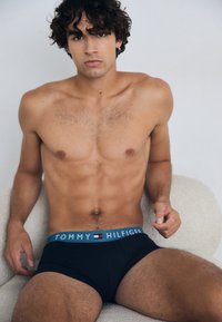 Un sous-vêtement noir avec une ceinture bleue portant le logo "TOMMY HILFIGER". Le modèle a des cheveux bouclés et est assis, dans un environnement de fond texturé de couleur claire.