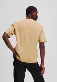 Beige rövidujjú póló puha anyagból, klasszikus kerek nyakkivágással és egyenes szabással, a szegélyénél kis logóval.