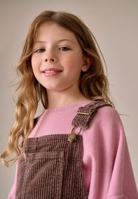 Brune corduroy overalls med sidelommer, parret med en lys pink sweater. Overallstropperne har gyldent hardware og tekstureret stof.