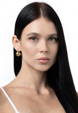Cercei din aur în formă de lacrimă, cu o suprafață lustruită și curbe subtile. Model cu păr lung negru și machiaj natural.