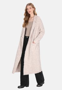 Donna che indossa un lungo cappotto beige texturizzato sopra un top taupe e pantaloni neri a gamba larga, in piedi con una mano nella tasca del cappotto.