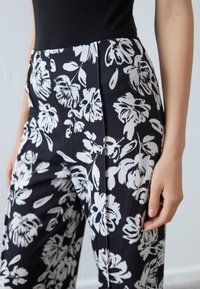 Pantalons noirs avec de grands motifs floraux blancs, ajustés, texture lisse et taille haute, vus de la taille au milieu de la cuisse.