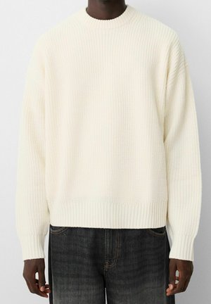 Mężczyzna ubrany w kremowy, dziergany sweter z prążkowanymi mankietami i dołem, w połączeniu z ciemnymi jeansami.