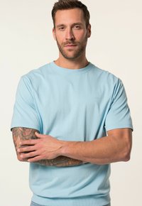 Světle modré bavlněné tričko s krátkými rukávy, kulatým výstřihem a decentním logem na hrudi. Model má tetování na pažích.