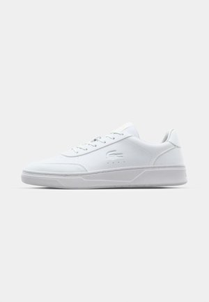 Hvit sneaker med en glatt syntetisk overdel, flate lisser, subtile perforeringer og en teksturert såle. Har en liten logo på siden.