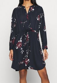 Dunkelblaues Kleid mit langen Ärmeln, Knöpfen vorne, Blumenmuster in Burgunderrot und Weiß sowie einem eingeschnürten Bund für eine verstellbare Passform.