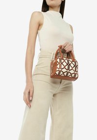 Bruine handtas met een geometrisch crèmekleurig patroon, voorzien van korte handvatten en een rits. Model houdt de tas vast en draagt een aansluitend beige topje en lichte broek.