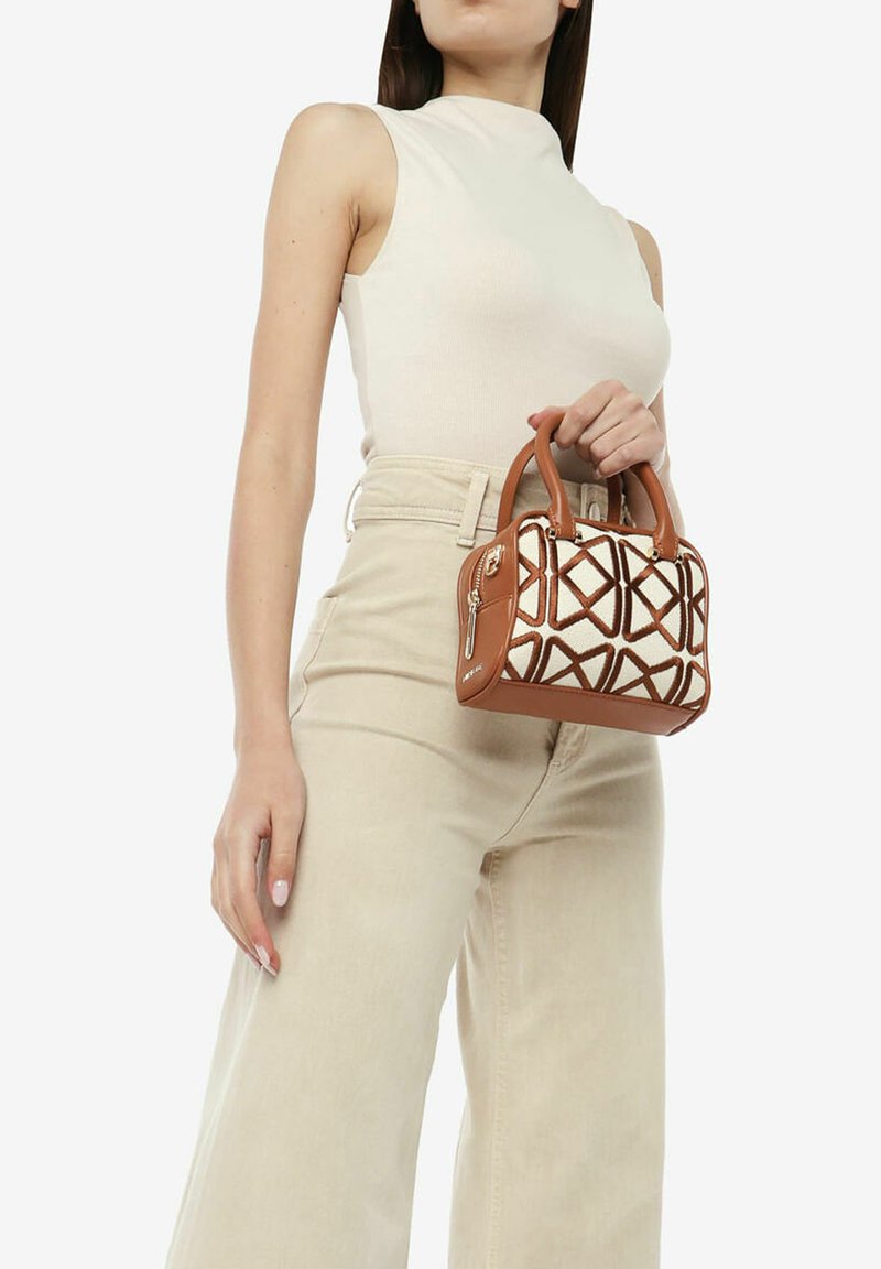 Bruine handtas met een geometrisch crèmekleurig patroon, voorzien van korte handvatten en een rits. Model houdt de tas vast en draagt een aansluitend beige topje en lichte broek.