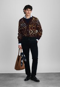 Cardigan tricoté en bordeaux avec des motifs géométriques multicolores, associé à un pull bleu marine, un pantalon noir, des chaussures noires et un sac fourre-tout marron.