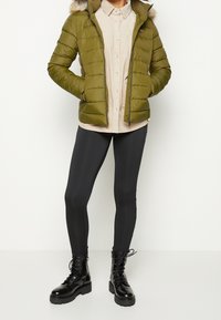 Person iført en olivengrøn polstret jakke med pelshætte, beige skjorte med knapper, sorte leggings og sorte snørebånds kampstøvler.