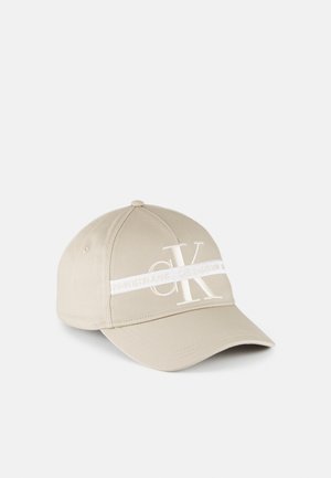 Casquette de baseball beige à visière courbée, avec de grandes lettres blanches "CK" et une bande blanche horizontale avec un petit texte sur le devant.