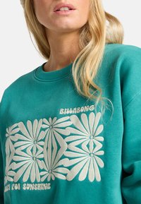 Felpa teal con grafica floreale bianca e testo ricamato "Billabong" e "Back for Sunshine." Texture morbida, design con collo a giro.