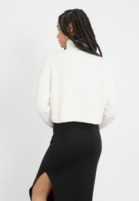 Gekürzter weißer Pullover mit hochgeschlossenem Kragen und geripptem Saum, kombiniert mit einem schwarzen Rock mit einem hochgeschnittenen Schlitz. Glatte Textur, schlichtes Design.
