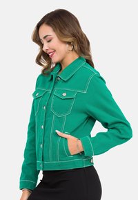 Grüne Jeansjacke mit zwei Vordertaschen, kontrastierendem weißen Nähten, Knopfverschluss und Kragen.
