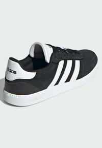 Zapatilla negra con diseño de tres franjas blancas, parte superior de ante, cuello acolchado y suela de goma blanca. Presenta detalles en el talón texturizados en negro.