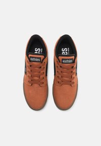 Brown canvas sneakers with black accents, round toe, lace-up design, rubber sole, and Etnies branding on the tongue and side.