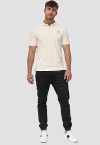 Cremfarbendes Poloshirt mit kurzen Ärmeln, drei-Knopf-Leiste und Logo. Kombiniert mit schwarzen, taillierten Jogginghosen und weißen Sneakern.
