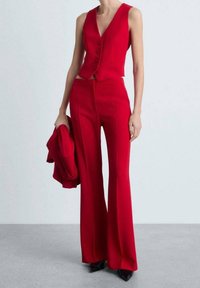 Rotes, maßgeschneidertes Weste und ausgestellte Hose, mit einem schicken Kragen und vorderen Knöpfen. Das Outfit wird mit einer passenden Jacke kombiniert, die in einer Hand gehalten wird.