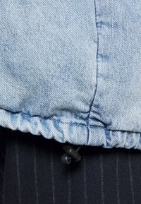 Gros plan sur une veste en denim bleu clair montrant un ourlet froncé avec un cordon de serrage ajustable, un tissu texturé et un fond à rayures noires contrastantes.