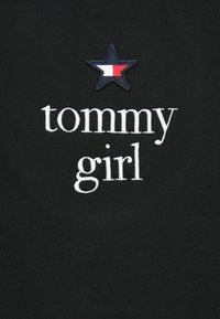 Čierna tkanina s bielym vyšívaným textom "tommy girl" pod námornou hviezdou vyšitou s bielymi a červenými časťami.