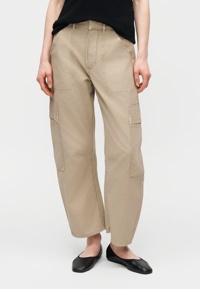 Γυναίκα που φοράει μπεζ παντελόνι cargo με φαρδιά μπατζάκια και τσέπες με επιθέματα, μαζί με μαύρα, επίπεδα παπούτσια, στέκεται μπροστά σε απλό φόντο.