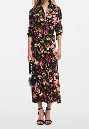 Woman wearing a long black floral dress with a belt, holding a small black handbag, and wearing black ankle-strap heels.