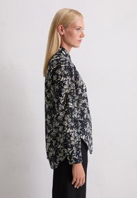 Femme blonde en blouse à manches longues noir à fleurs et pantalon noir, standing de profil contre un mur blanc uni.