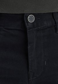Los jeans de mezclilla negra cuentan con un botón metálico visible con detalle de logo, costuras en contraste y una textura suave. La parte superior tiene una cinturilla de canalé verde.