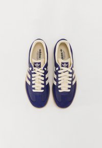 Tmavě modré tenisky s krémovými detaily, bílé tkaničky, design se třemi pruhy, polstrovaný límec a texturovaná gumová podešev.