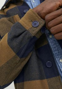 Ruskea ja laivastonsininen ruudullinen napitettava paita, jossa on flanellimainen rakenne, pyöreät laivastonsiniset napit ja denimpaita kerroksittain alla.