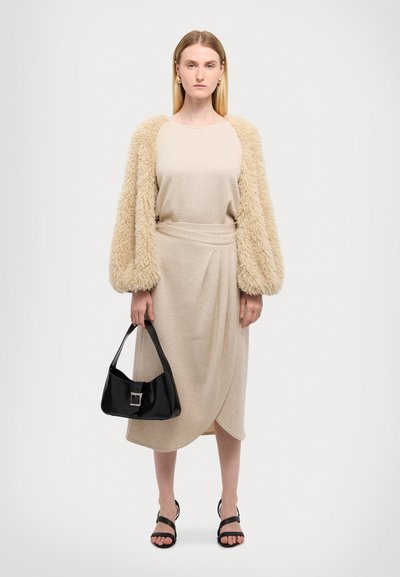 Beiges gestricktes Kleid mit asymmetrischem Saum, darunter eine flauschige beige Strickjacke. Schwarze Tasche mit silbernen Beschlägen und schwarze Riemchensandalen.