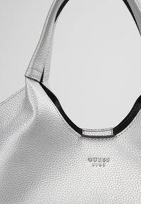 Zilveren faux leren handtas met een gestructureerde afwerking, voorzien van een zwarte rand en een opvallend logo aan de voorkant.