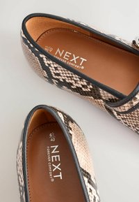 Teksturoitu, keinonahkainen kävelykenkä mustan ja vaaleanpunaisen sävyissä, jossa on ruskea pohjallinen, jossa lukee brändi "NEXT FOREVER COMFORT."