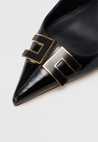 Μαύρο παπούτσι από δέρμα βινυλίου με μυτερή μύτη, που διαθέτει μια μεγάλη μαύρη και χρυσή γεωμετρική αγκράφα στο μπροστινό μέρος. Λείο υφή με ελάχιστες ραφές.