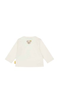 Chemise crème à manches longues avec un col rond, présentant un patch brodé d'ours au dos et une étiquette jaune à l'ourlet. Tissu en coton doux.