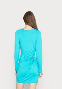 Vestido de manga longa em turquesa com uma silhueta ajustada, detalhe franzido na lateral e um recorte acima da bainha. Tecido suave e elástico.