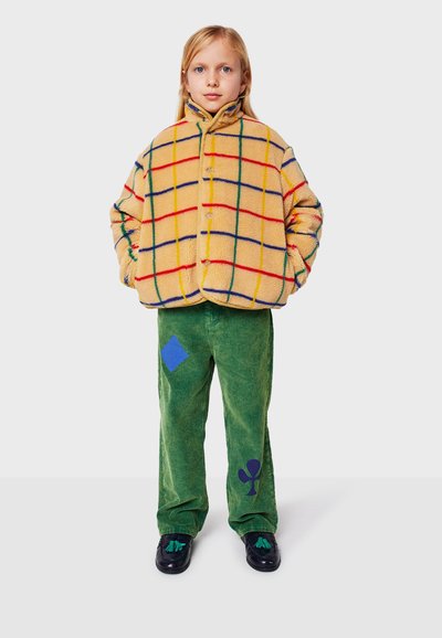 Giacca in pile a scacchi con linee gialle, rosse e blu; pantaloni in cotone verde a coste con forme di diamanti e fiori blu; scarpe nere.