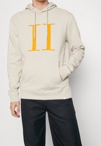 Sudadera crema con un gran gráfico frontal naranja "II". Presenta una capucha con cordón, un bolsillo tipo canguro y puños acanalados.