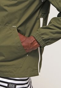 Ciemnoskóra dłoń w zielonej oliwkowej kieszeni kurtki z białym zamkiem błyskawicznym, na czarnych i białych spodniach w paski z widoczną etykietą BKN.
