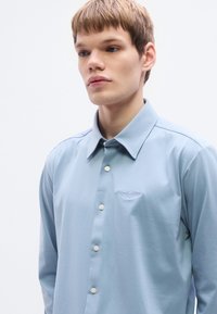Φωτεινό γαλάζιο πουκάμισο με μακριά μανίκια από λείο υλικό, με μυτερό γιακά και κεντημένο λογότυπο στο στήθος, κλείνει με κουμπιά.