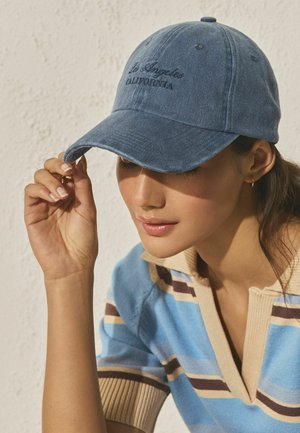 Woman adjusting blue cap embroidered with "Los Angeles California," wearing a striped blue and beige short-sleeve top against a textured wall.