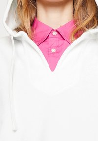 Bílá mikina s designem do V a se šňůrkami, nošená přes jasně růžovou košilovou halenku s knoflíkovými detaily a hladkou texturou.