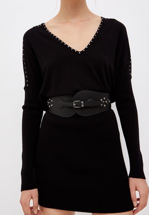 Vrouw draagt een zwarte lange mouwen jurk met V-hals en zilverkleurige studs langs de halslijn en mouwen, gecombineerd met een brede zwarte riem met gesp.