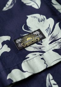 Tejido azul marino con patrones florales en verde claro; presenta una etiqueta negra con texto dorado y un gráfico de sol que dice "Superdry."