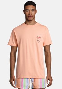 Camiseta de algodón en melocotón claro con mangas cortas y escote redondo. Presenta un gráfico colorido en el lado izquierdo del pecho.