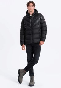 Schwarze Steppjacke mit Kapuze und Zickzack-Quiltmuster, ausgestattet mit zwei Reißverschlusstaschen. Getragen mit schwarzen Jeans und grauen Schnürstiefeln.