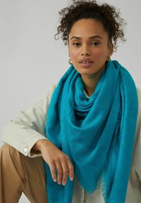 Écharpe turquoise avec un motif texturé et des bords à franges, drapée autour du cou. Chemise de couleur claire partiellement visible en arrière-plan.
